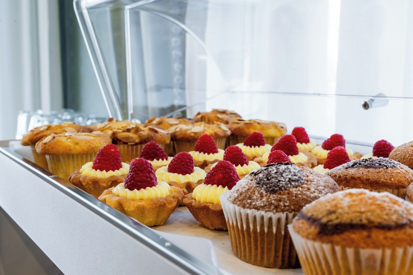 Una teca con diversi prodotti da forno, tra cui muffin e pasticcini, alcuni guarniti con lamponi.
