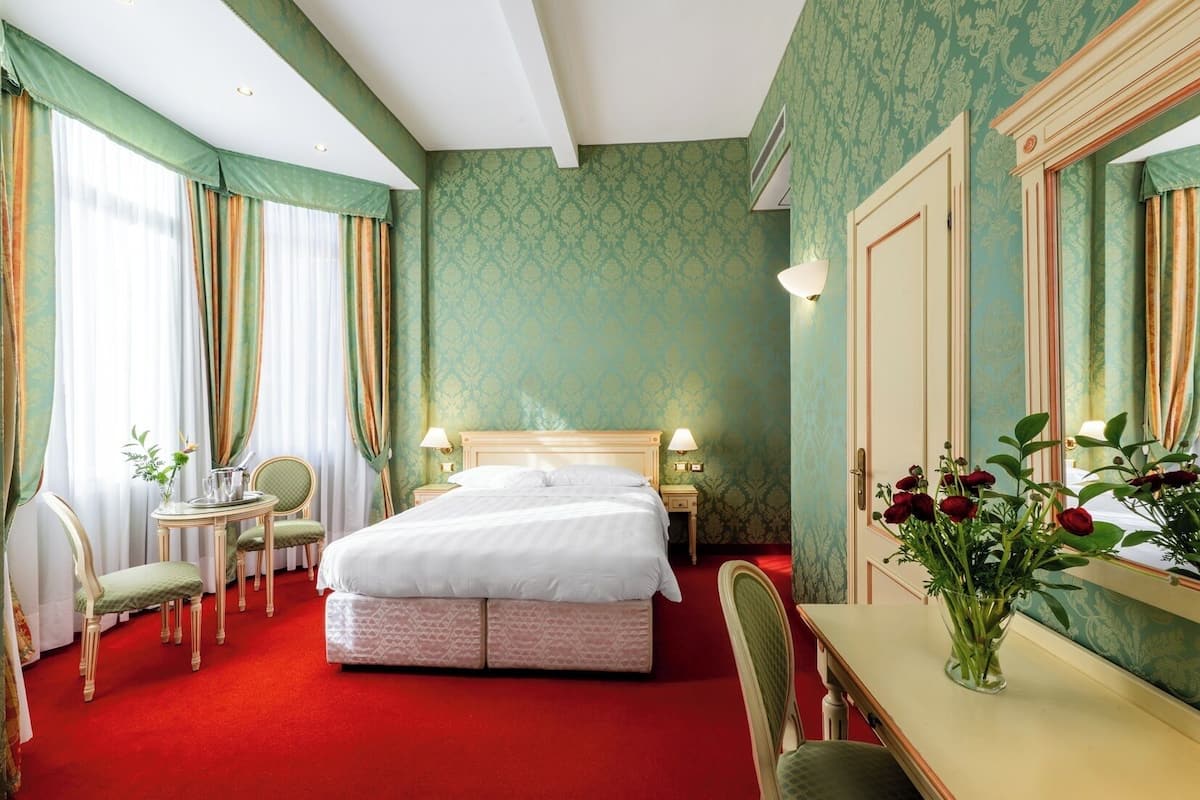 Une chambre d’hôtel avec un grand lit, deux tables de chevet, un bureau, une chaise, une petite table ornée d’un vase de fleurs et un miroir.