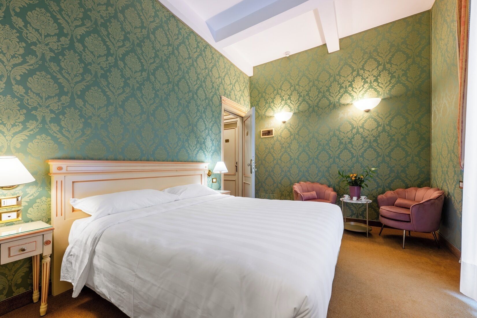 A hotel room with a large bed, two chairs, a small table, and a nightstand with a lamp.