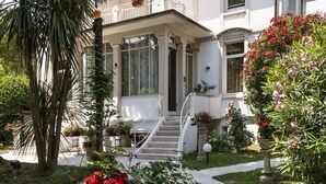 Um edifício branco com varanda coberta, escadaria e jardim com diversas plantas e flores.