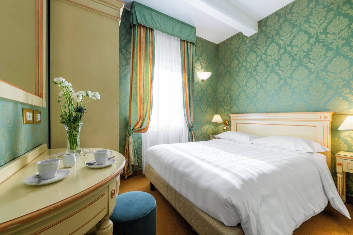 Une chambre d’hôtel avec un lit, une coiffeuse avec un miroir, un vase de fleurs et une fenêtre avec des rideaux.