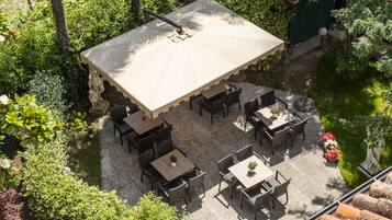Ein Terrassenbereich im Freien mit mehreren Tischen und Stühlen, umgeben von üppiger Bepflanzung und einem kleinen Wasserspiel.