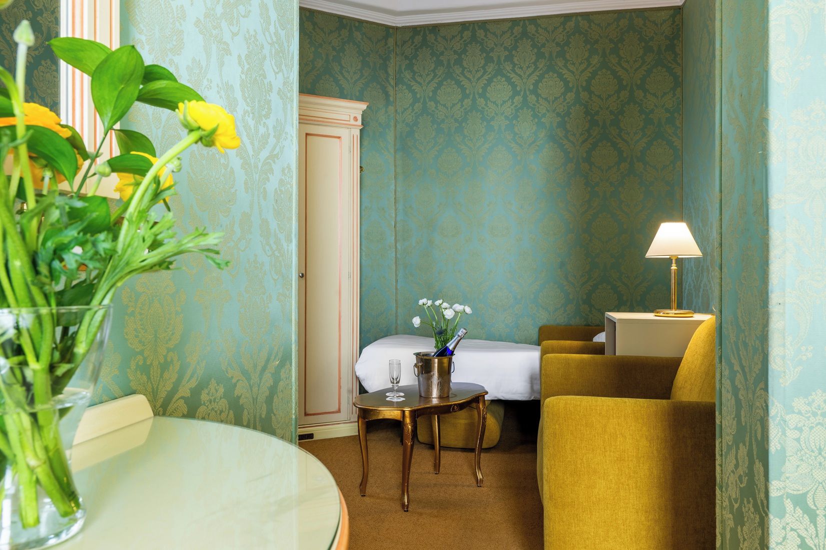 Quarto de hotel com cama, uma mesa pequena com abajur, uma cadeira e um vaso com flores.