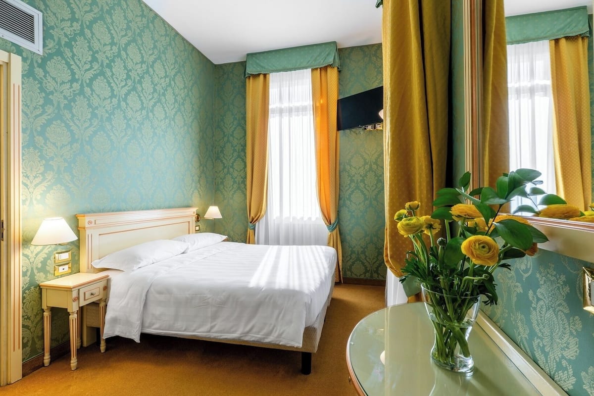 Une chambre d’hôtel avec un lit, des tables de chevet, un miroir et un vase de fleurs.