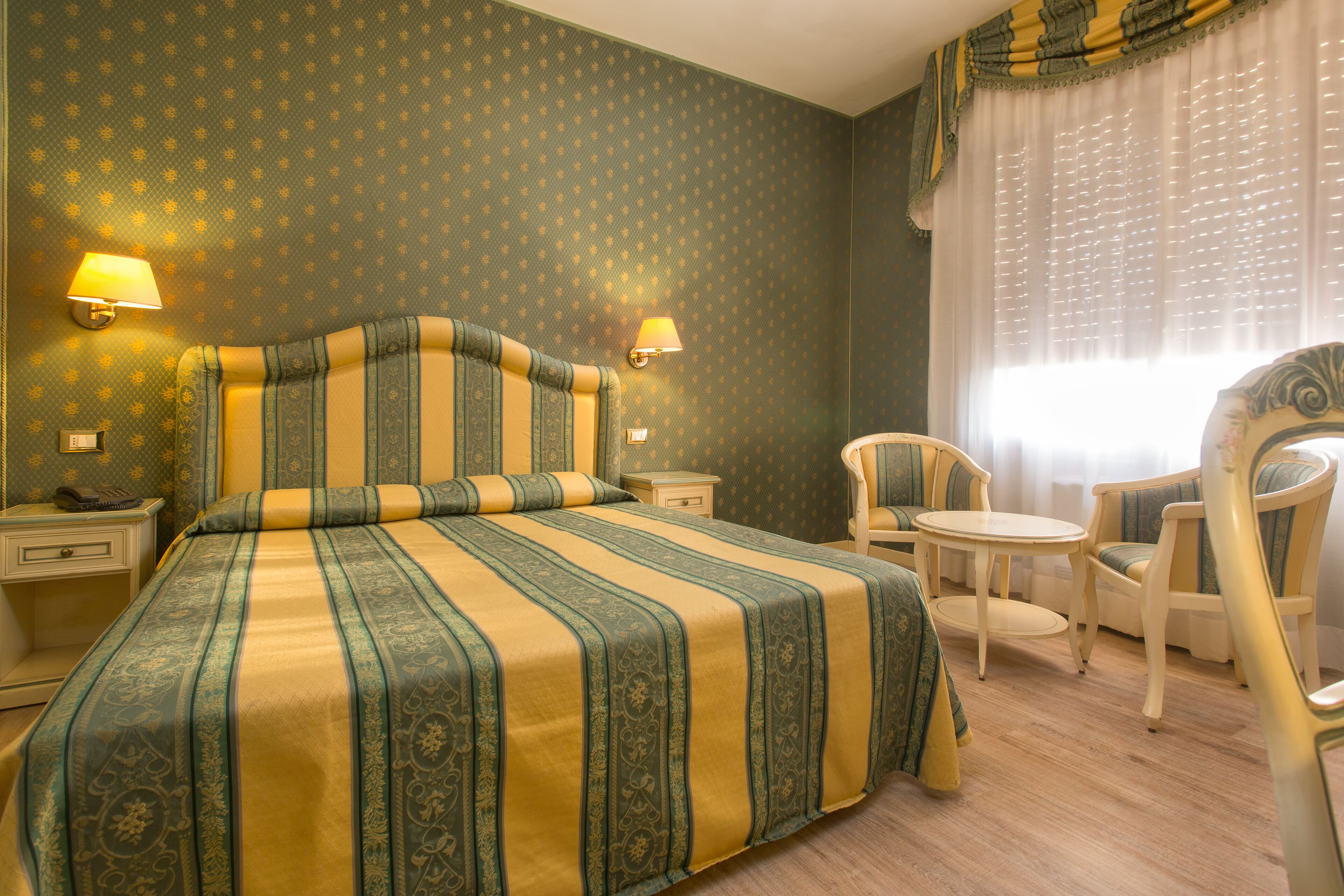 Ein Hotelzimmer mit einem großen Bett, zwei Nachttischen, einem Sessel, einem kleinen runden Tisch und einem Fenster mit Vorhängen.