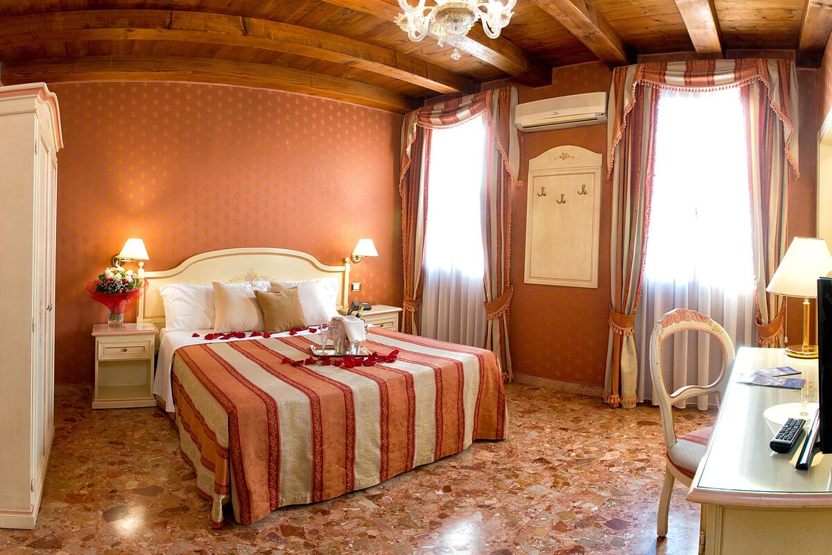 Una habitación de hotel con una cama grande, dos mesitas de noche con lámparas, un escritorio con una silla y una araña.