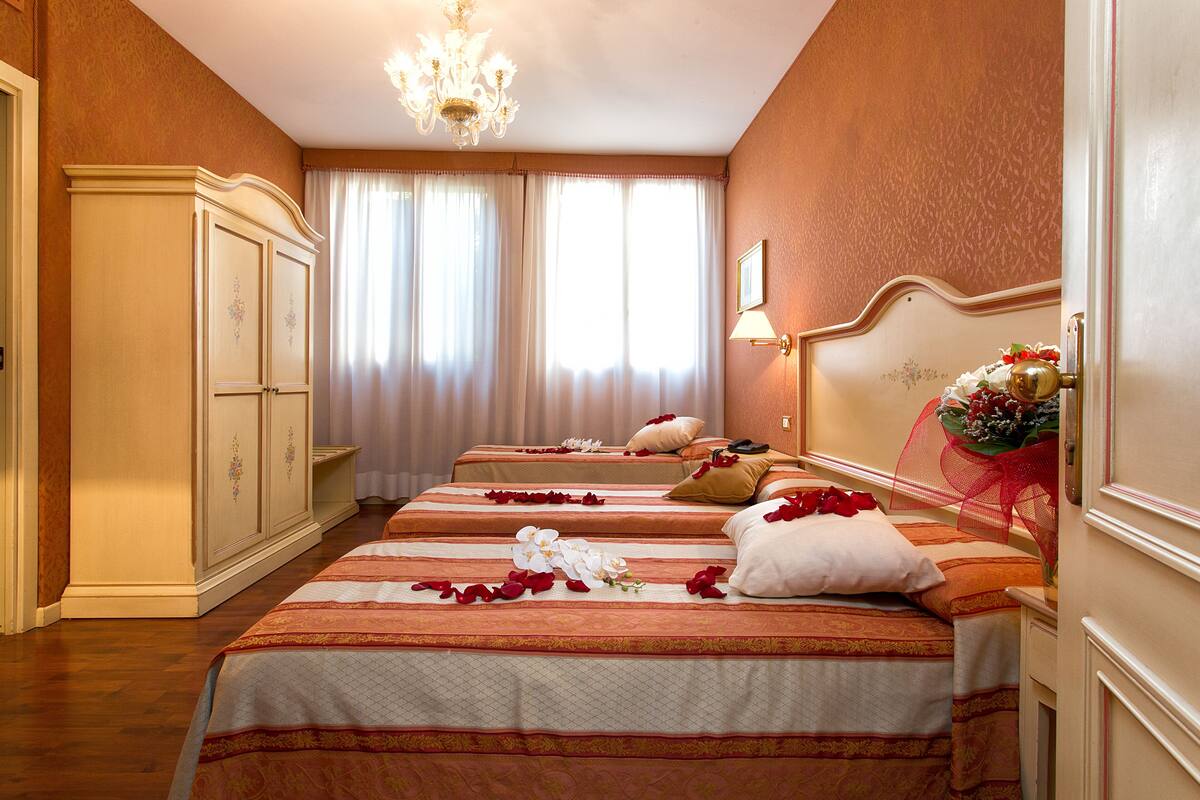 Habitación de hotel con dos camas, un armario, una araña y una ventana con cortinas.
