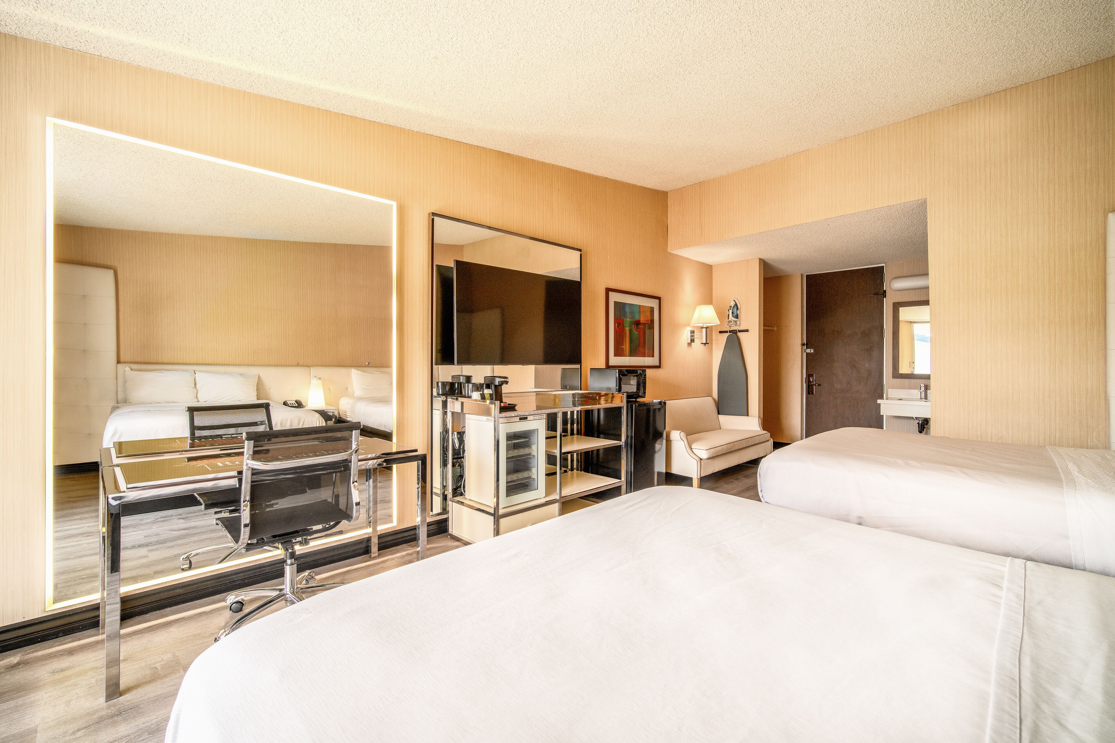 A hotel room with two beds, a desk, a mirror, a small kitchenette, and a bathroom.