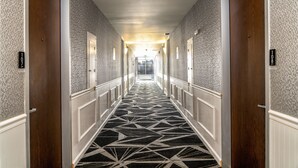 Een hotelgang met een geometrisch patroon in het tapijt, houten deuren en een lichtgekleurde muur.