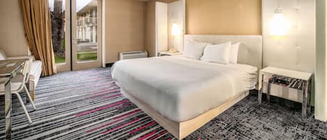 Quarto de hotel com uma cama grande, uma escrivaninha com cadeira, mesa de cabeceira, abajur e vista para o exterior.