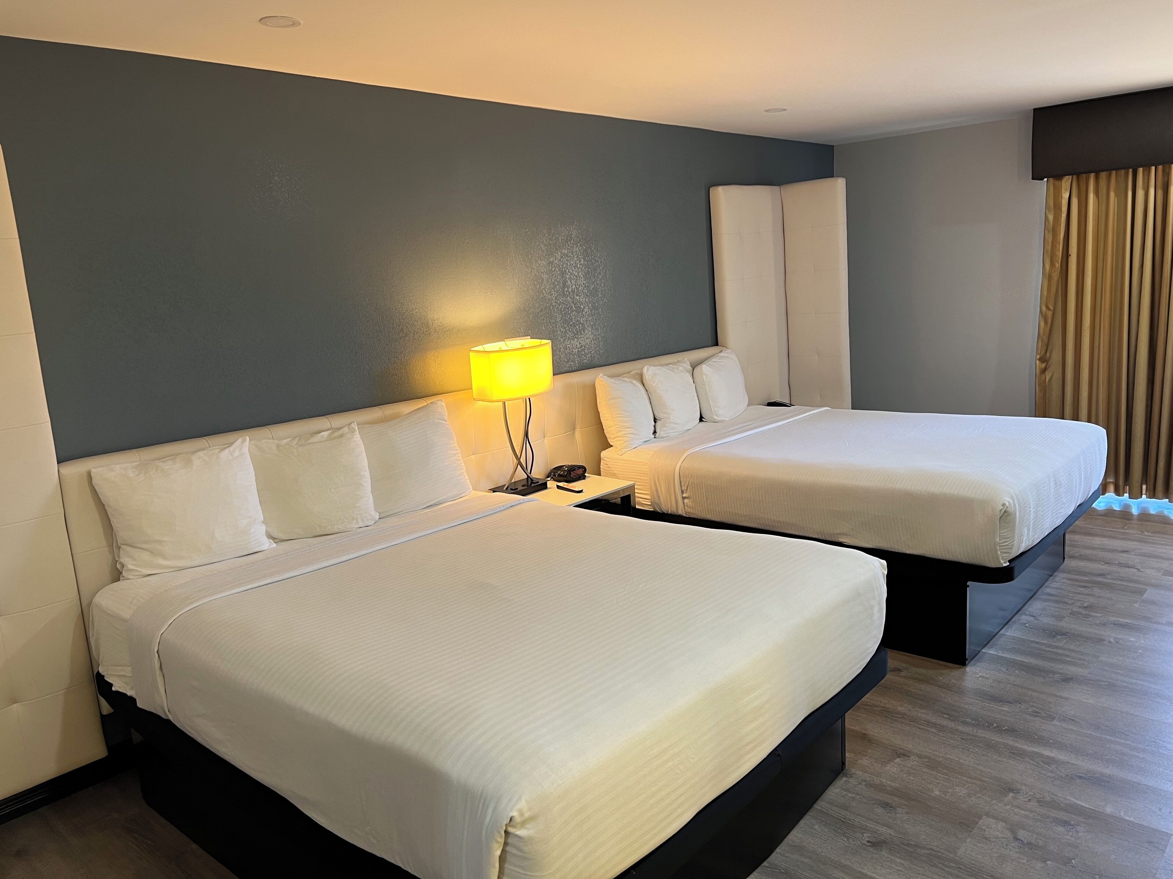 Hotellihuone, jossa on kaksi sänkyä, yöpöytä lampun kanssa ja suuri ikkuna, jossa on verhot.