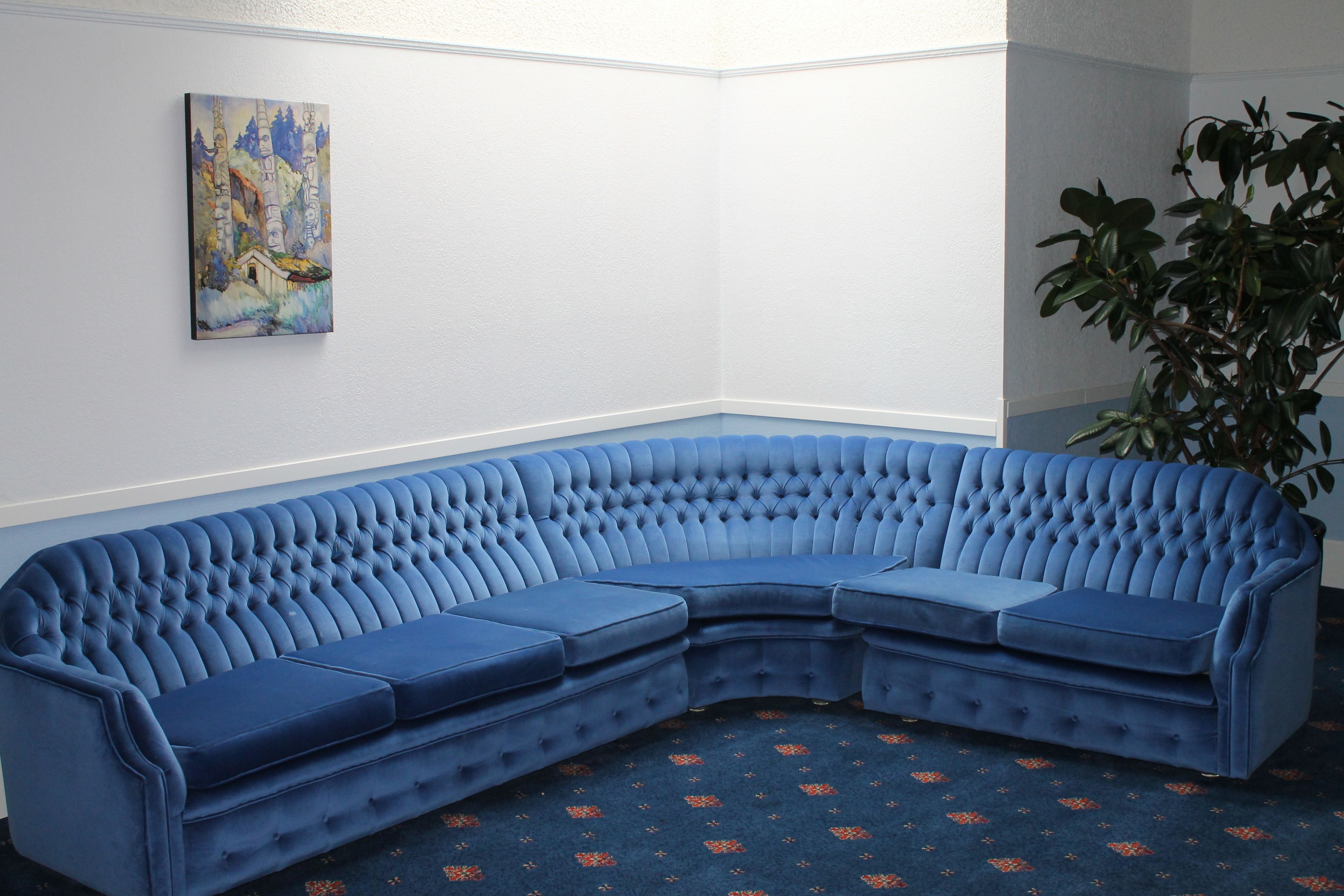 A corner room with a blue sectional sofa, a painting on the wall, and a potted plant.