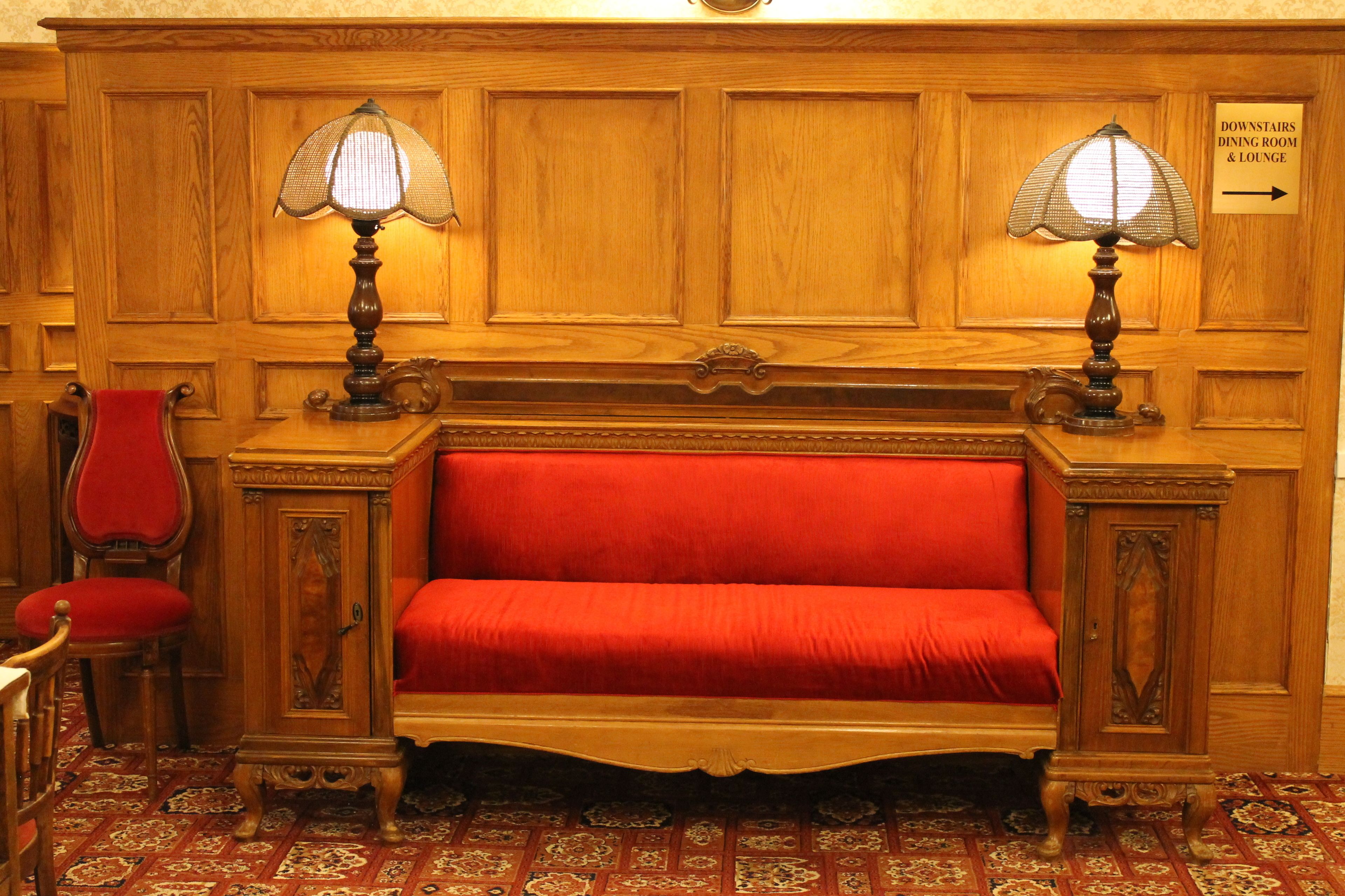 Un banco de madera con un cojín rojo, dos lámparas y un cartel que indica la ubicación del comedor y el salón en la planta baja.