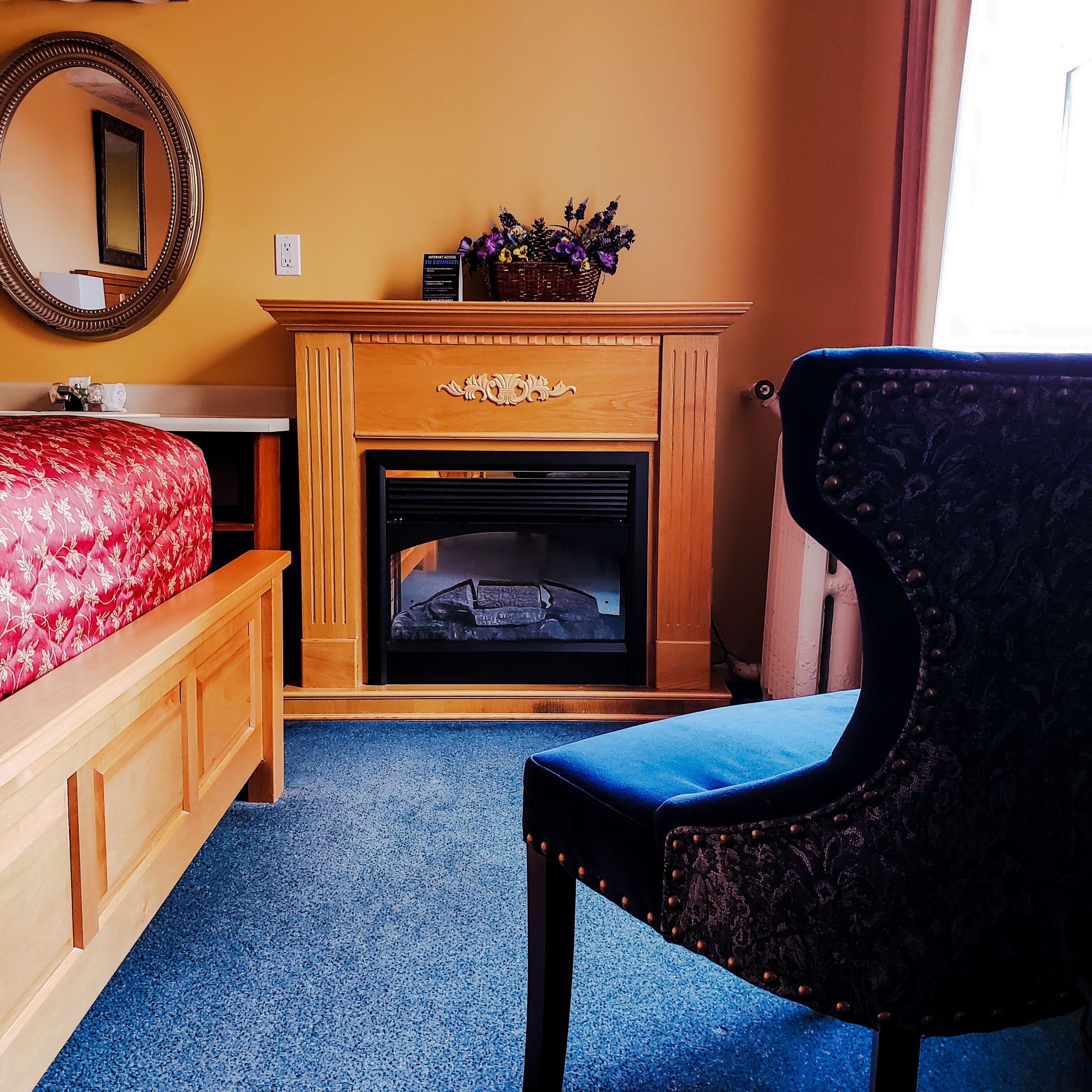 A bedroom with a wooden dresser, a red upholstered bed, a blue carpet, a black upholstered chair, a fireplace, and a mirror on the wall.