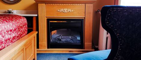 A bedroom with a wooden dresser, a red upholstered bed, a blue carpet, a black upholstered chair, a fireplace, and a mirror on the wall.