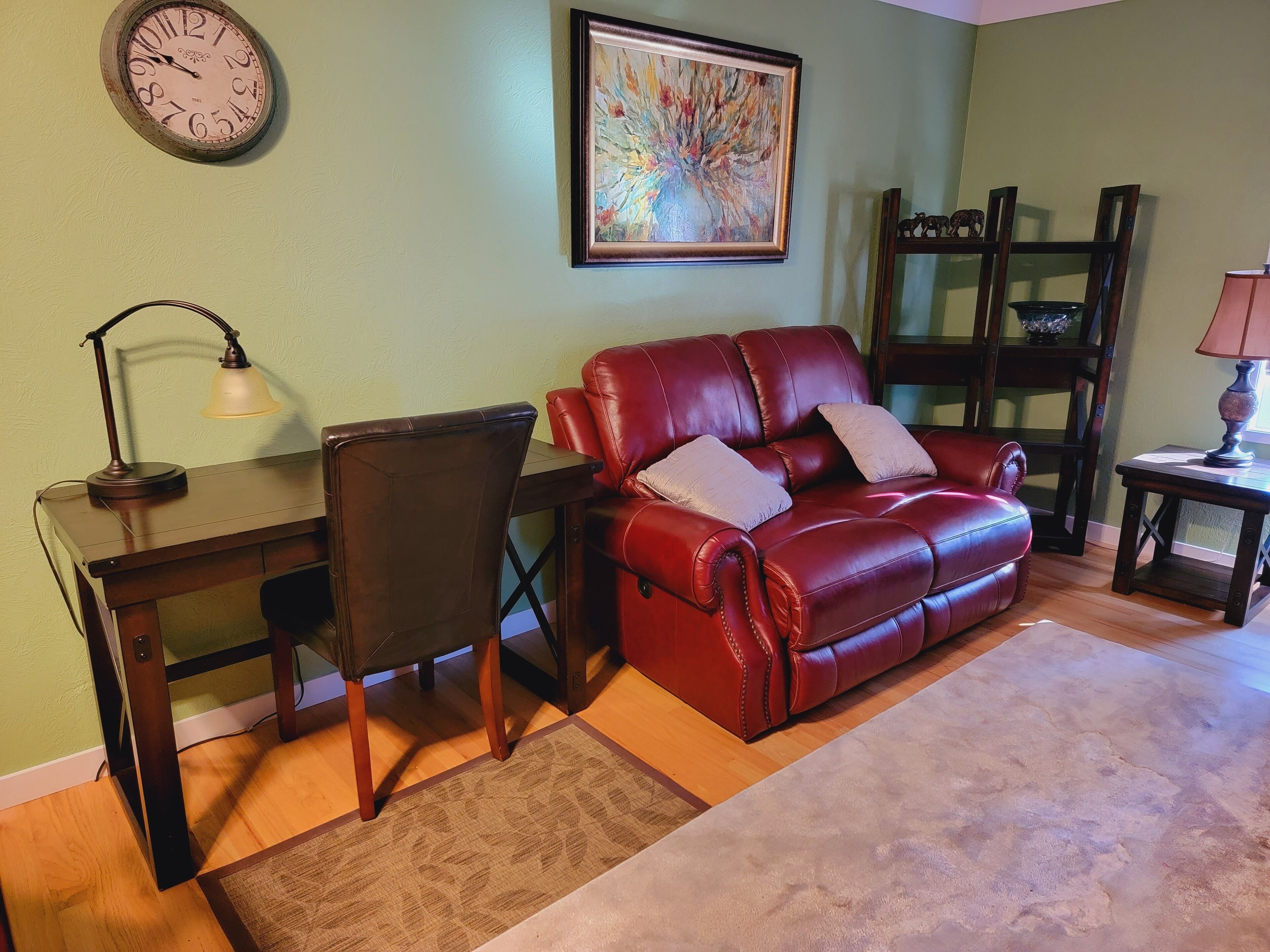A living room with a red leather sofa, a wooden desk, a chair, a lamp, a clock, and a framed abstract painting on the wall.