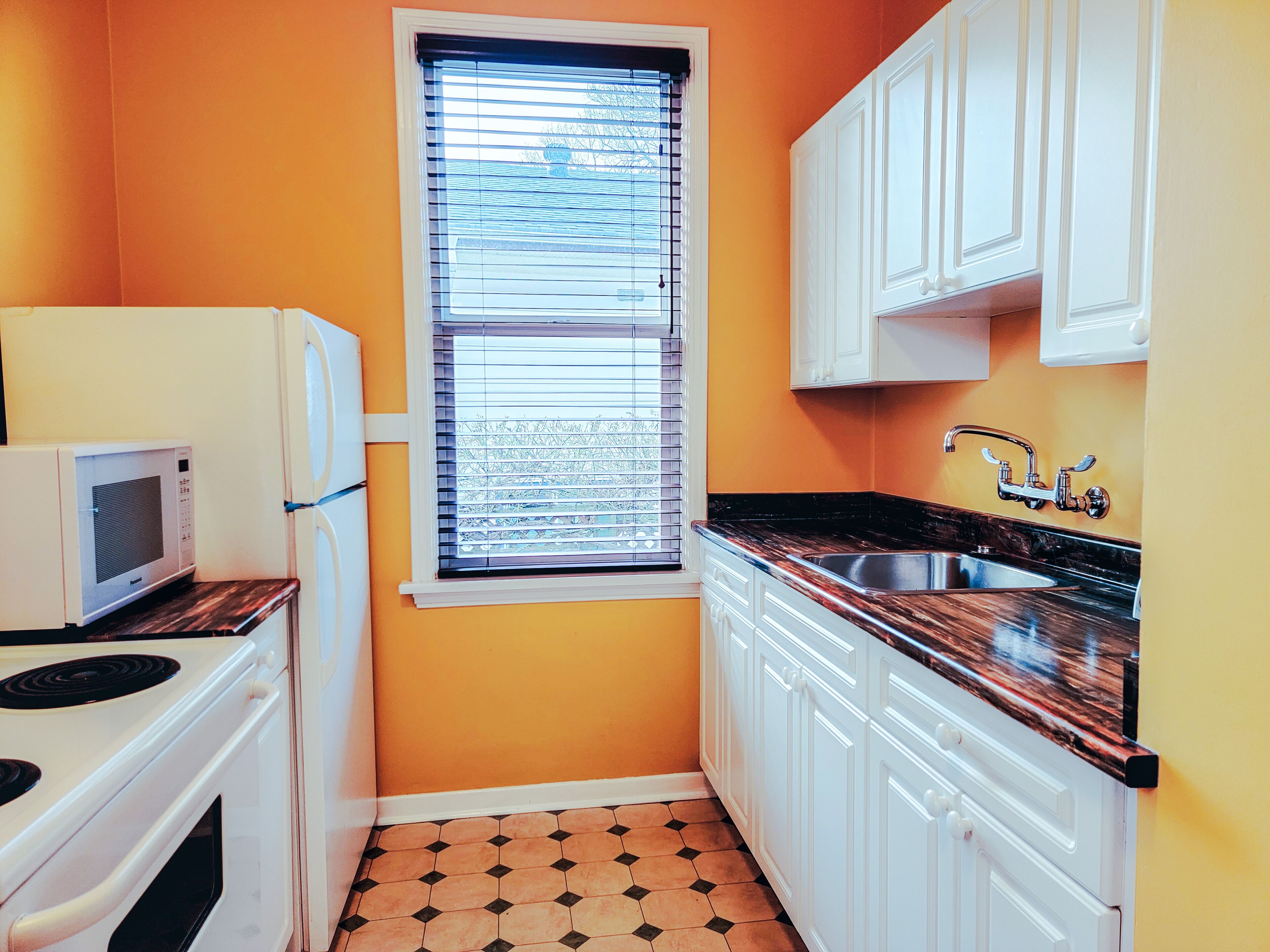 A compact kitchen with white cabinets, a black countertop, a window with blinds, and a microwave.