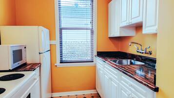 A compact kitchen with white cabinets, a black countertop, a window with blinds, and a microwave.
