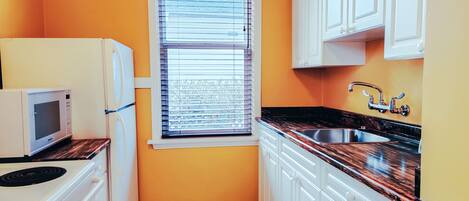 A compact kitchen with white cabinets, a black countertop, a window with blinds, and a microwave.