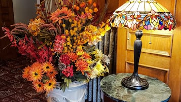 Eine weiße Keramikvase mit einer Blumenkomposition auf einem gemusterten Teppich, eine Holztür und eine Tischlampe mit einer Buntglaslampe.