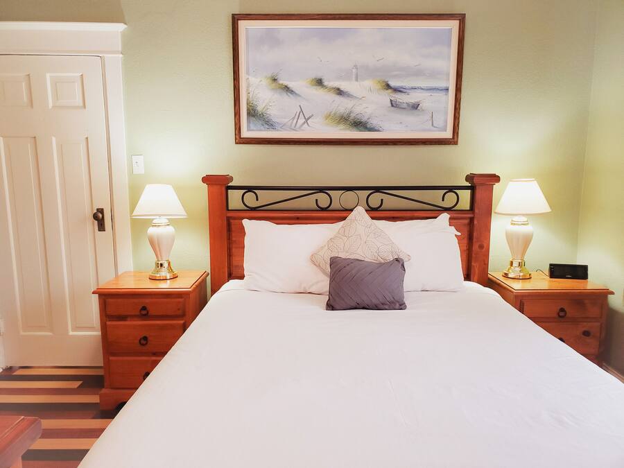 A neatly made bed with white linens and two pillows, flanked by wooden nightstands with lamps. A framed landscape painting hangs above the bed.