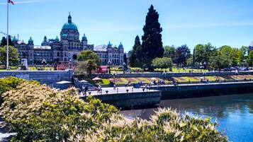 A large historic building with domed architecture, a river in the foreground, and a park with people and trees.