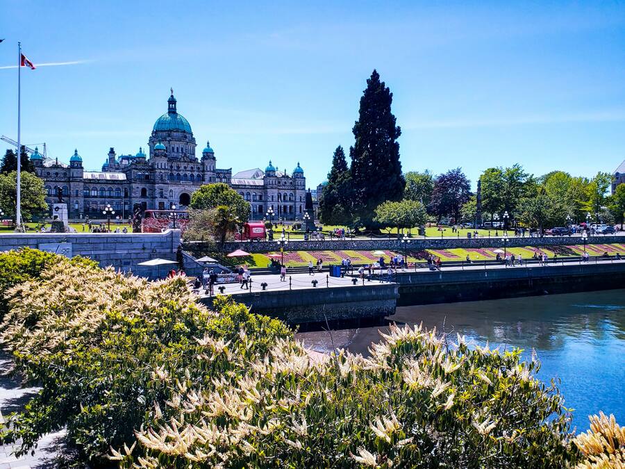 A large historic building with domed architecture, a river in the foreground, and a park with people and trees.