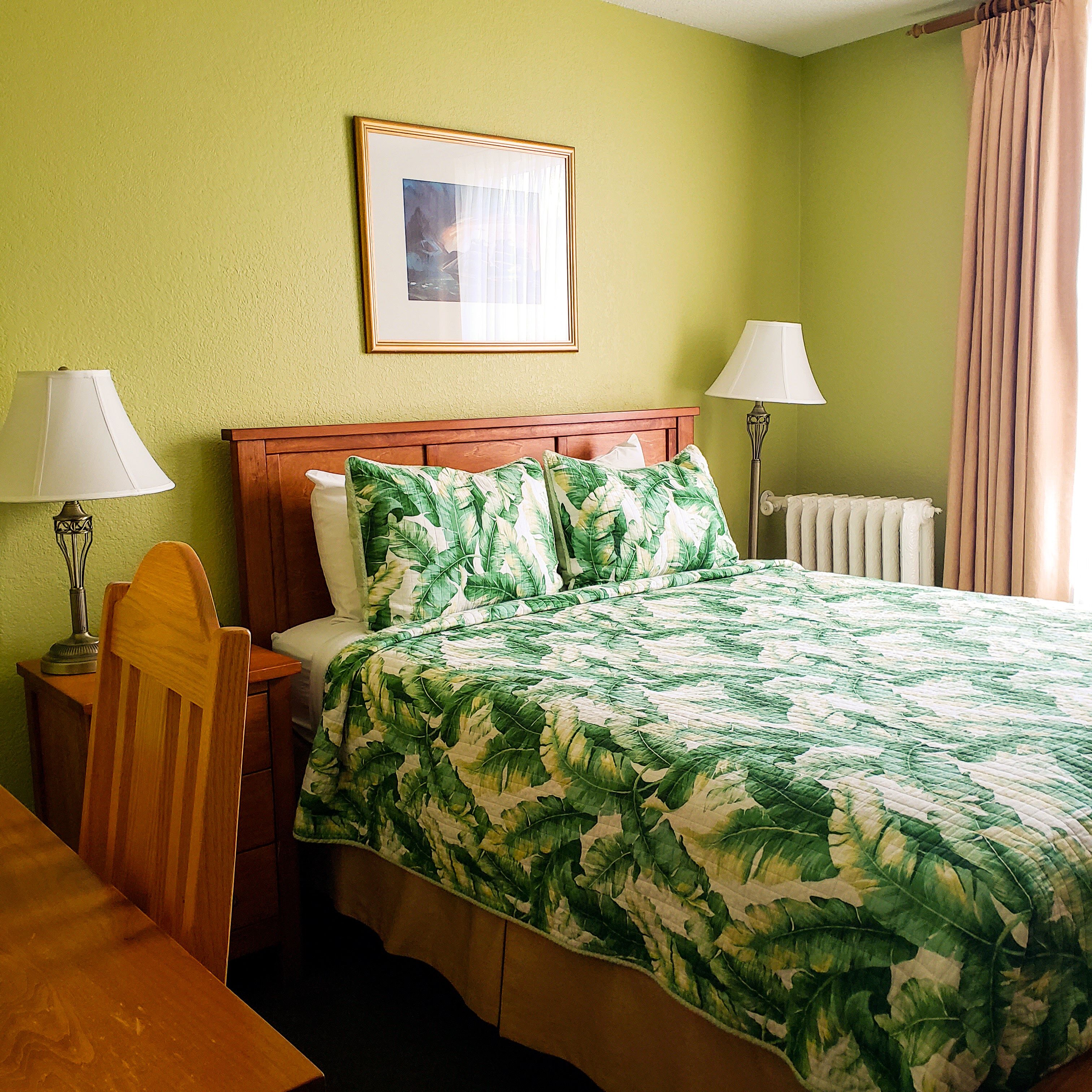 Habitación de hotel con una cama, dos lámparas de noche, una mesita de noche de madera, una silla, un cuadro en la pared y una ventana con cortinas.