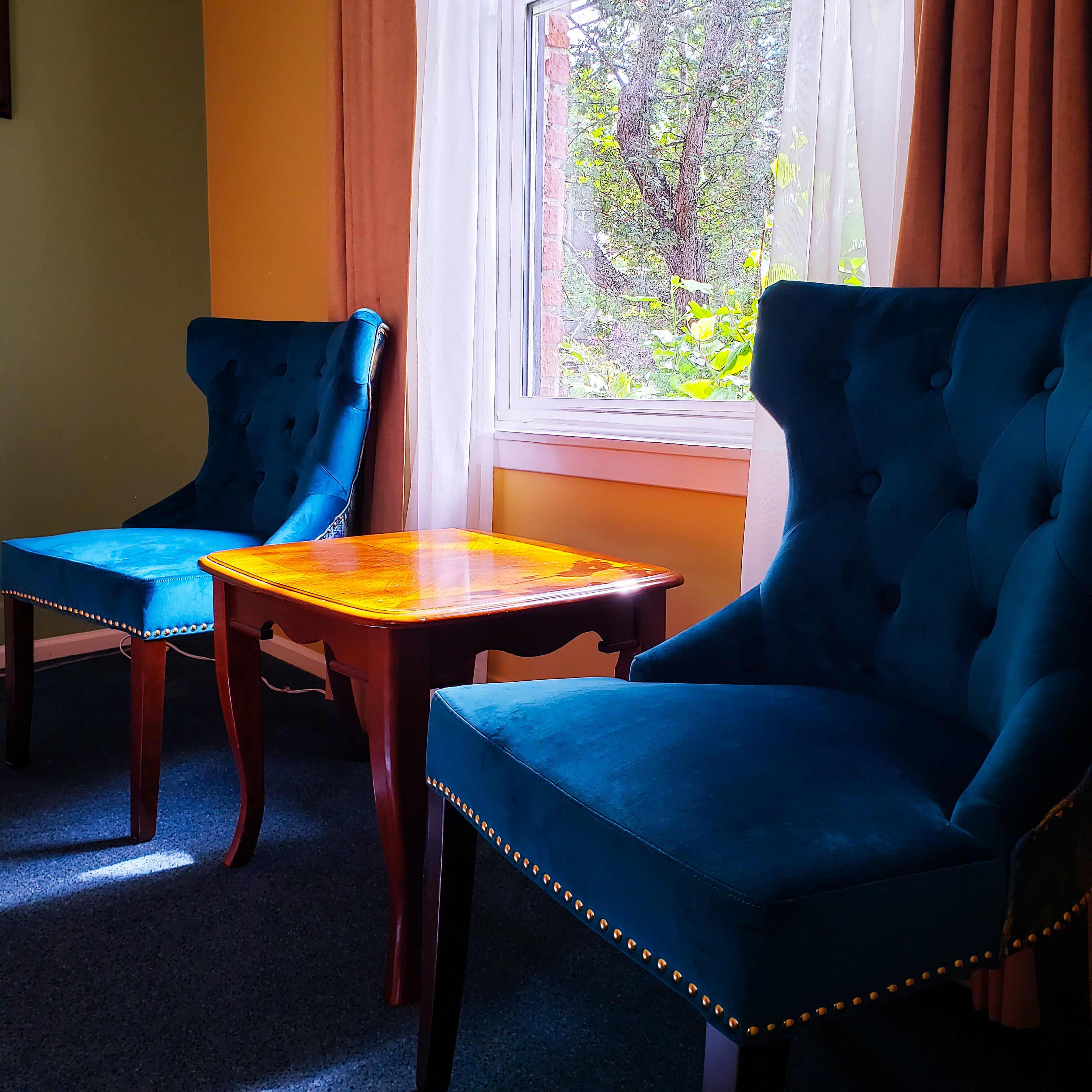 A room with two blue upholstered chairs, a wooden table, and a window with curtains.
