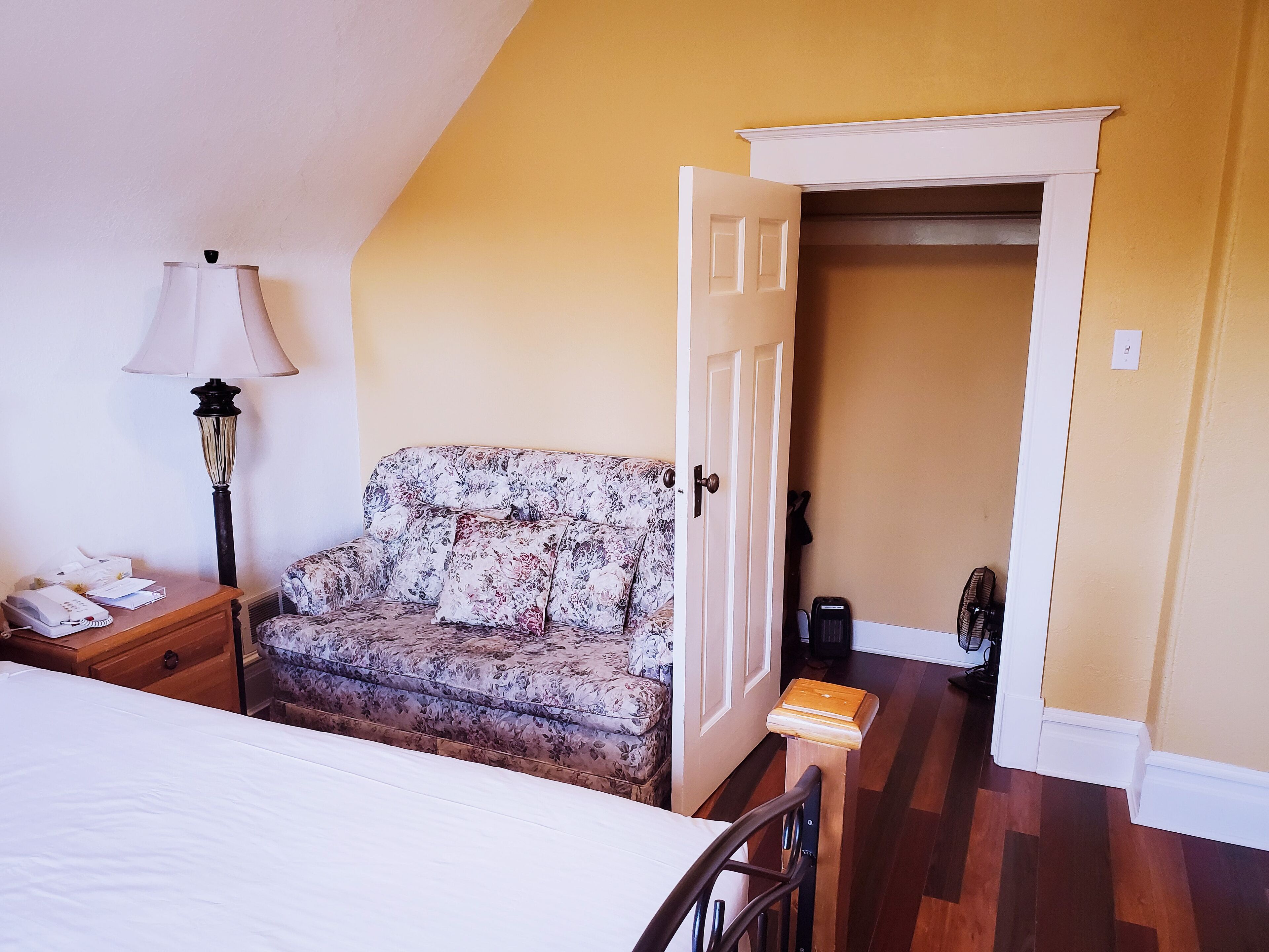 Quarto com cama, mesa de cabeceira, luminária, sofá floral, banco de madeira e porta que dá para outro cômodo.