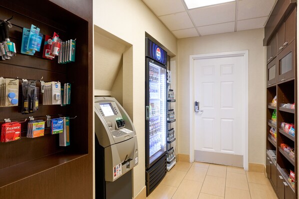 A hotel lobby with a self-service kiosk, a refrigerator stocked with beverages, and shelves with toiletries.