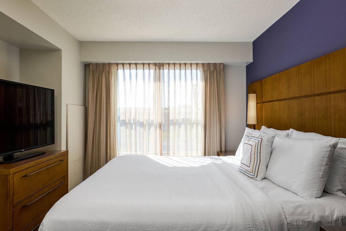 A hotel room with a large bed, a flat-screen TV on a wooden dresser, and a window with sheer curtains.