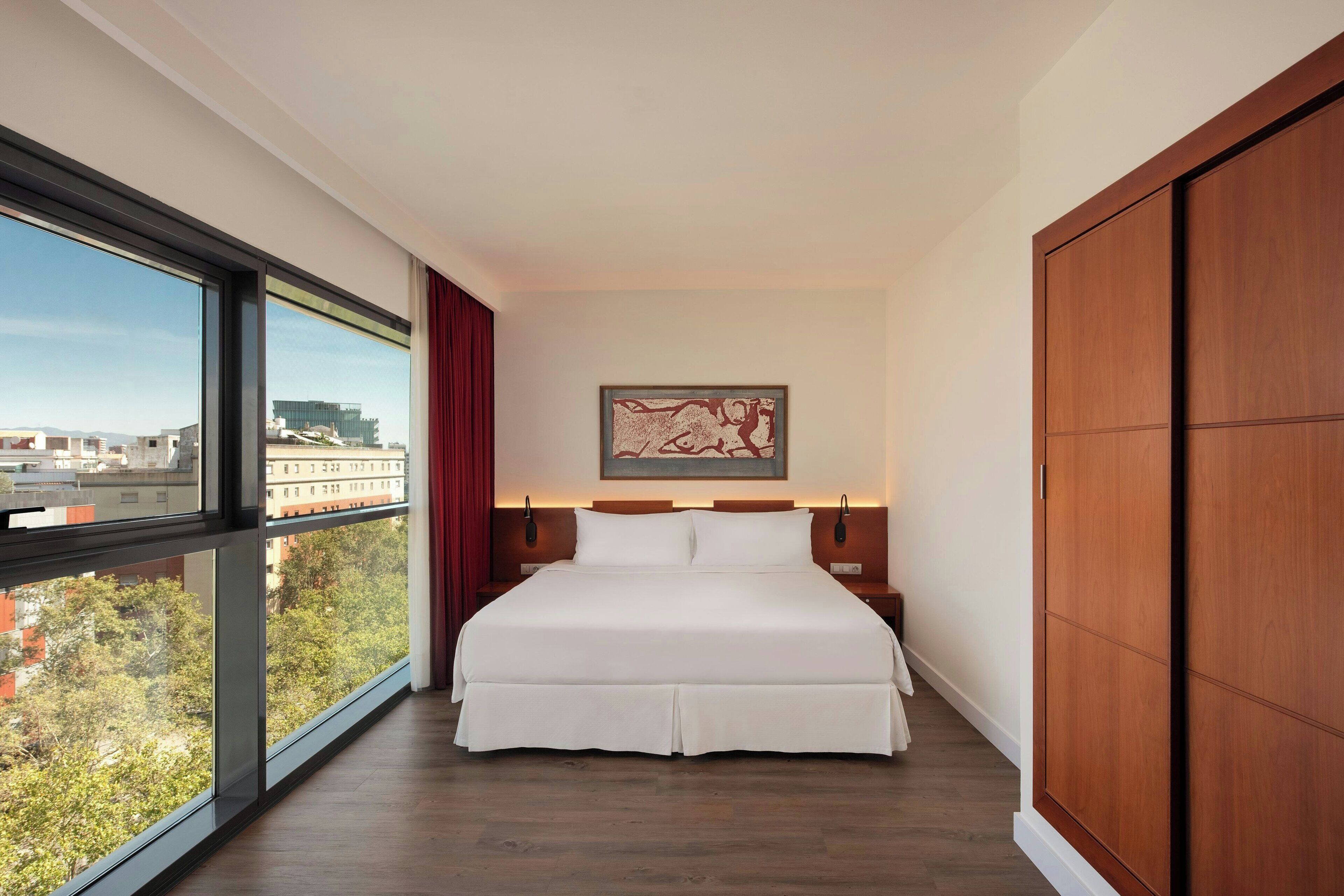 A modern hotel room with a large bed, a view through a large window, a wooden wardrobe, and a framed artwork on the wall.