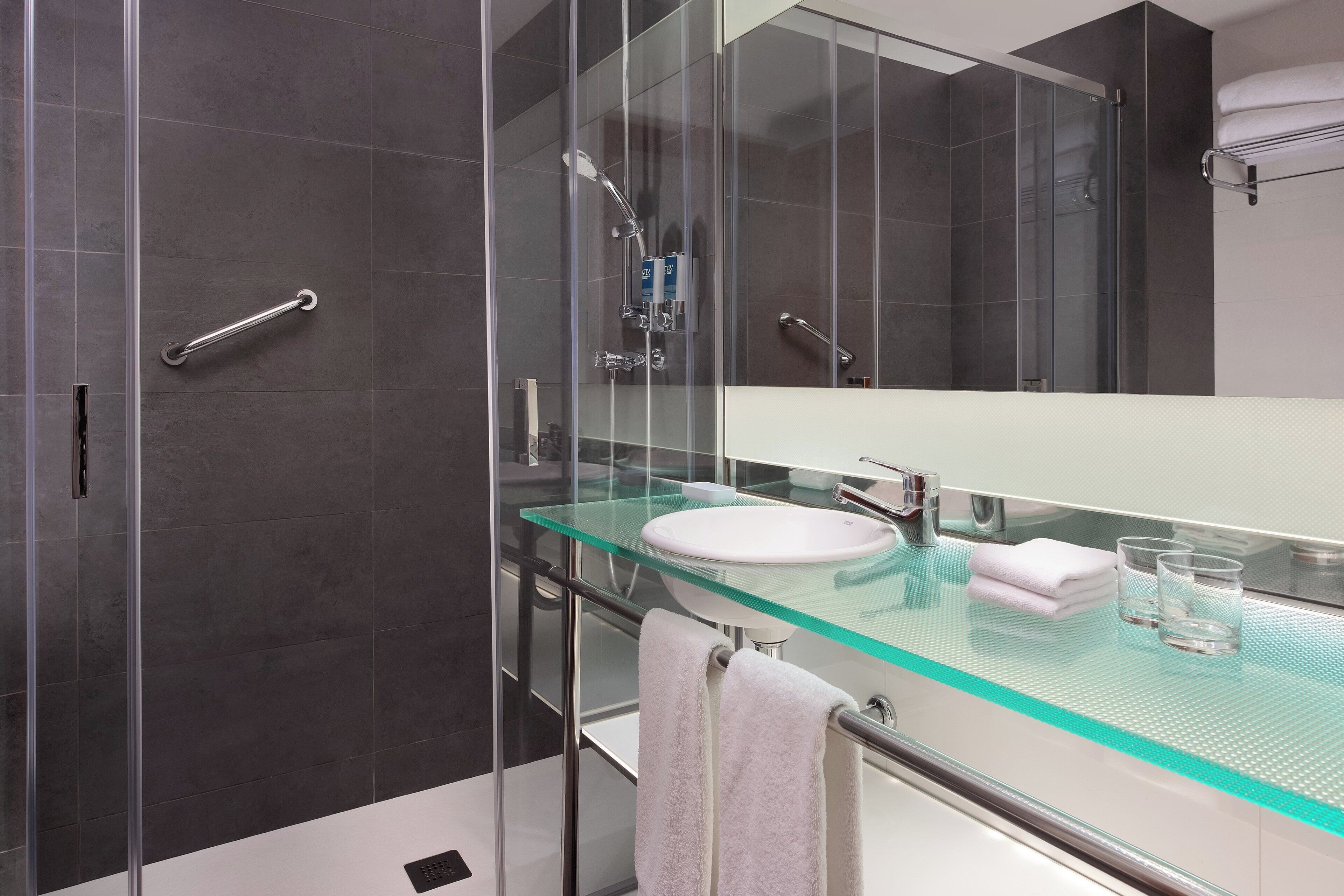 Une salle de bain moderne avec un lavabo en verre, des murs recouverts de carreaux foncés, une cabine de douche en verre et un porte-serviettes.