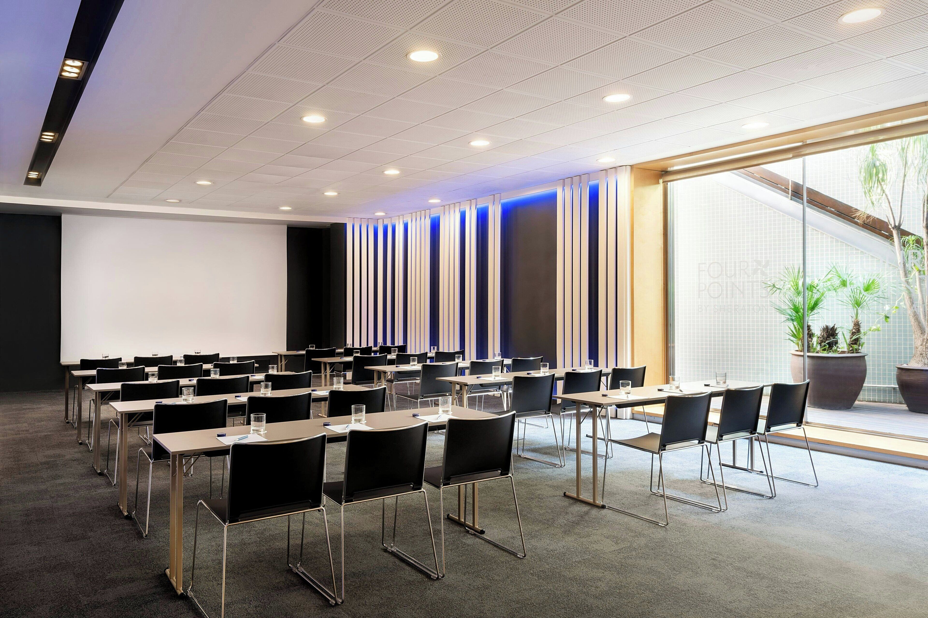 Une salle de conférence avec des rangées de tables et de chaises, un grand écran et une vue sur la verdure à l’extérieur.