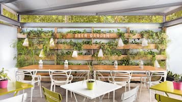 Un área de estar al aire libre acogedora, con una mesa y sillas de madera, rodeada de abundante vegetación y un letrero que dice “BEER GARDEN”.