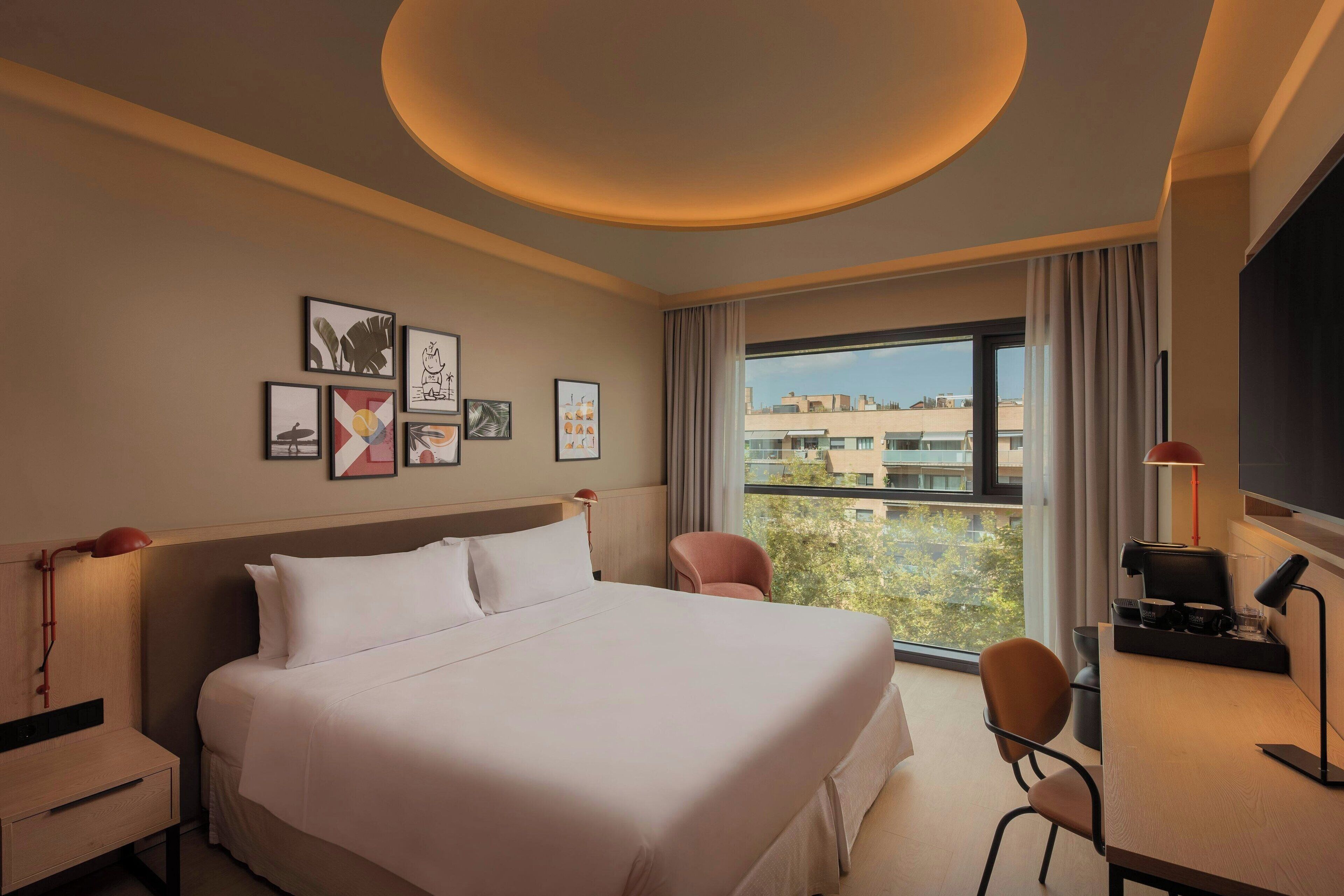 Habitación de hotel con una cama grande, un escritorio con silla, televisor y ventana con vistas a edificios.