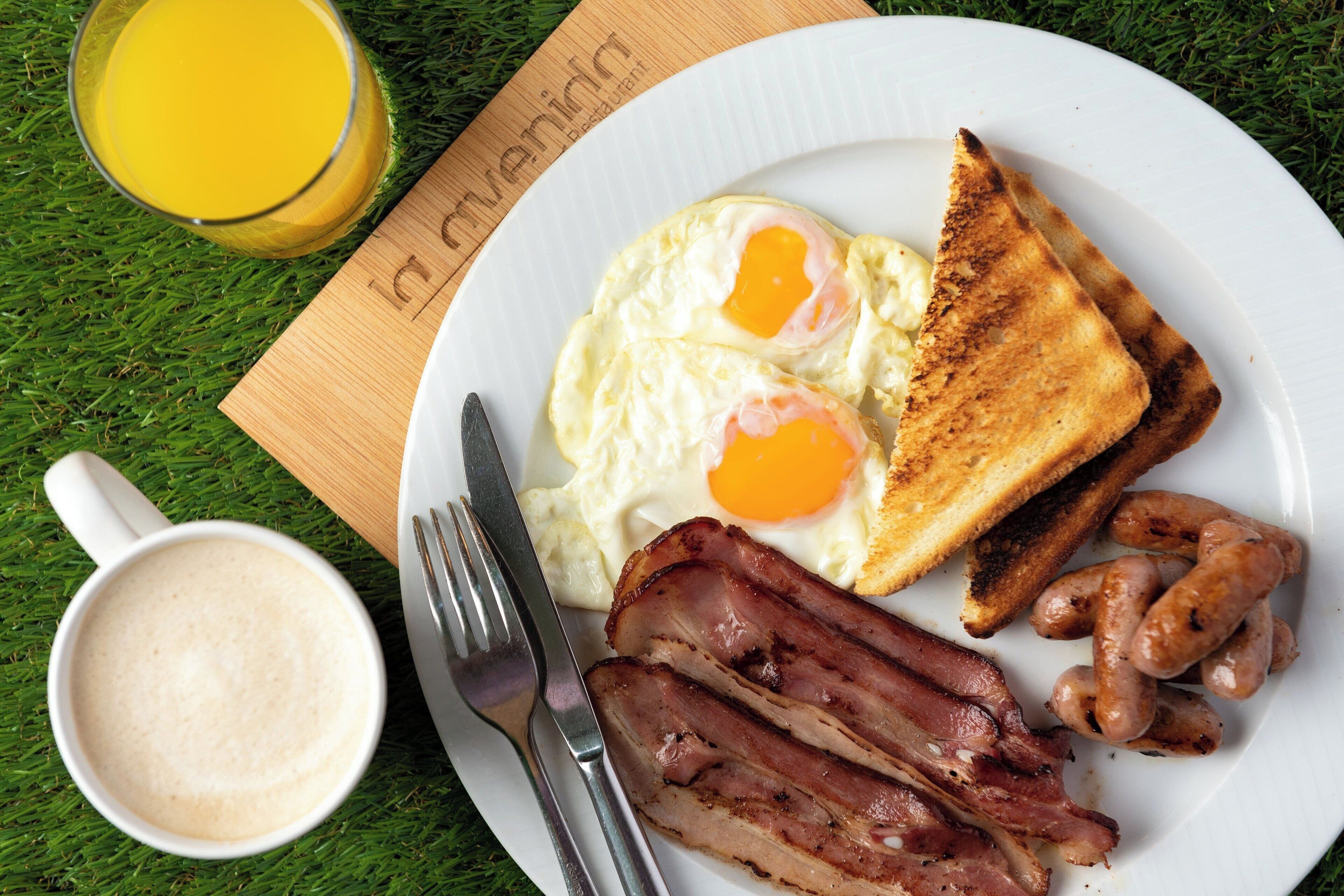 Un plato de desayuno con huevos, tostadas, tocino, salchichas y un vaso de jugo de naranja sobre un plato blanco.