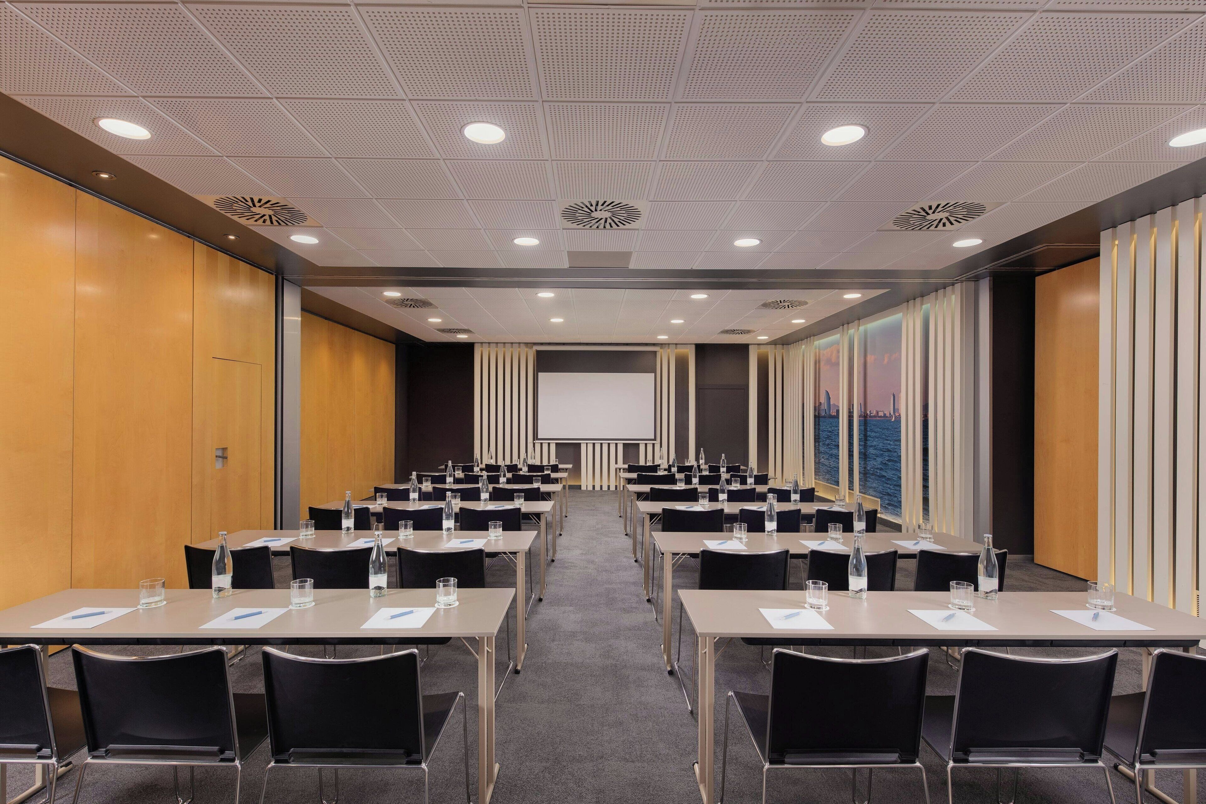 A conference room with rows of tables and chairs, a large screen, and a view of the sea through large windows.