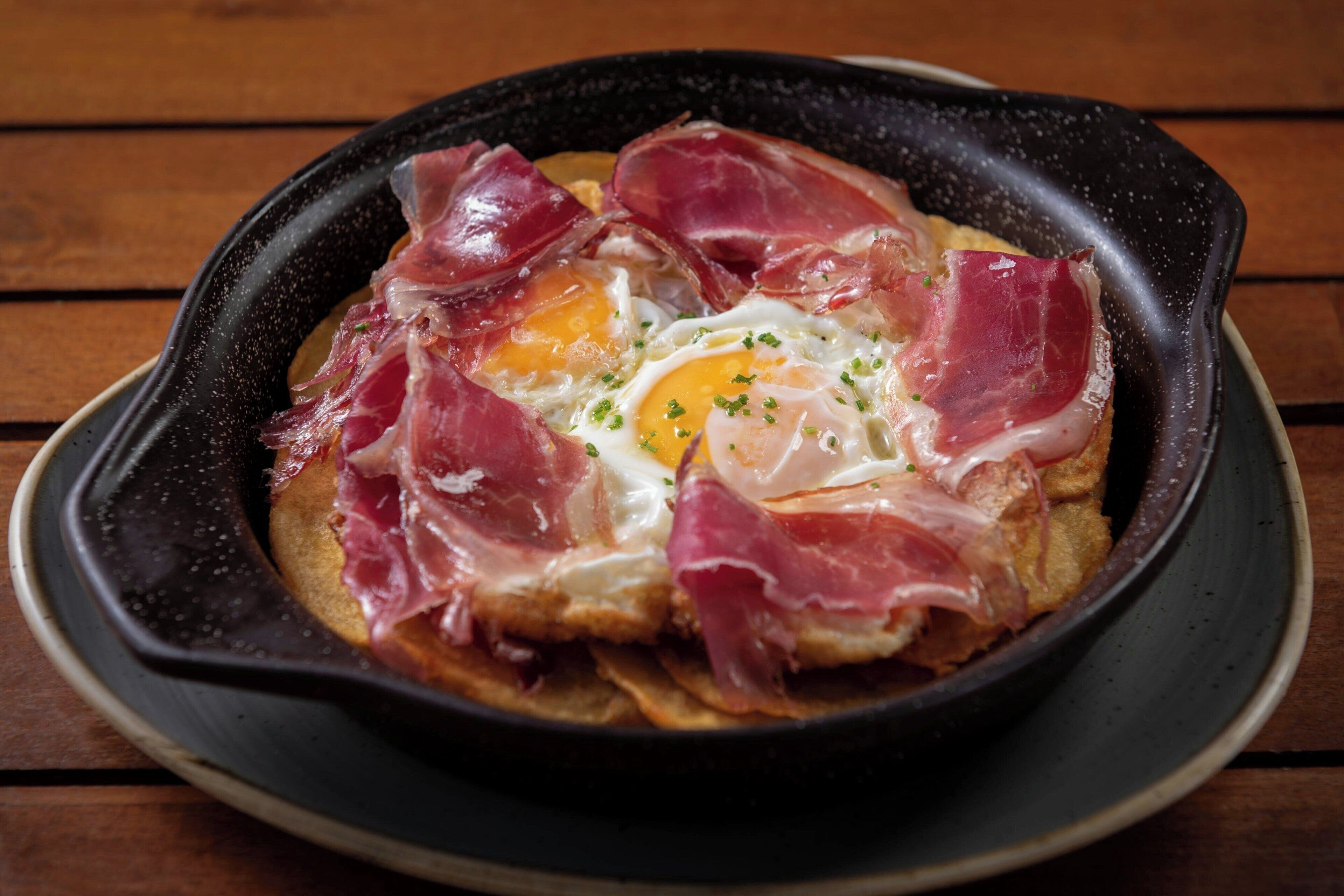 A dish with eggs and prosciutto on toast, served in a black skillet on a wooden table.