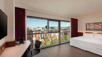 A hotel room with a large window, a bed, a desk with a chair, and a flat-screen TV.