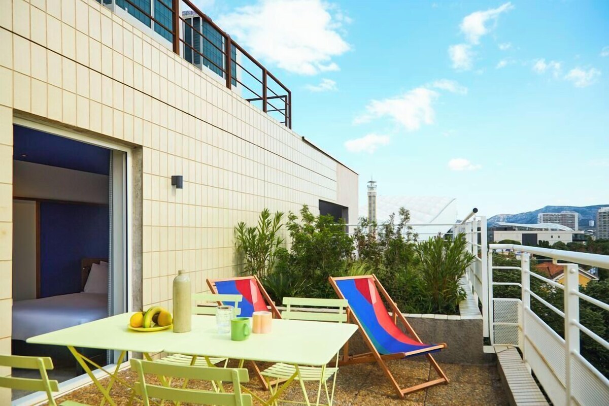 Een modern gebouw met een betegelde gevel, een balkon met groene stoelen en een tafel, en uitzicht op de stadsbelangrijkste punten.