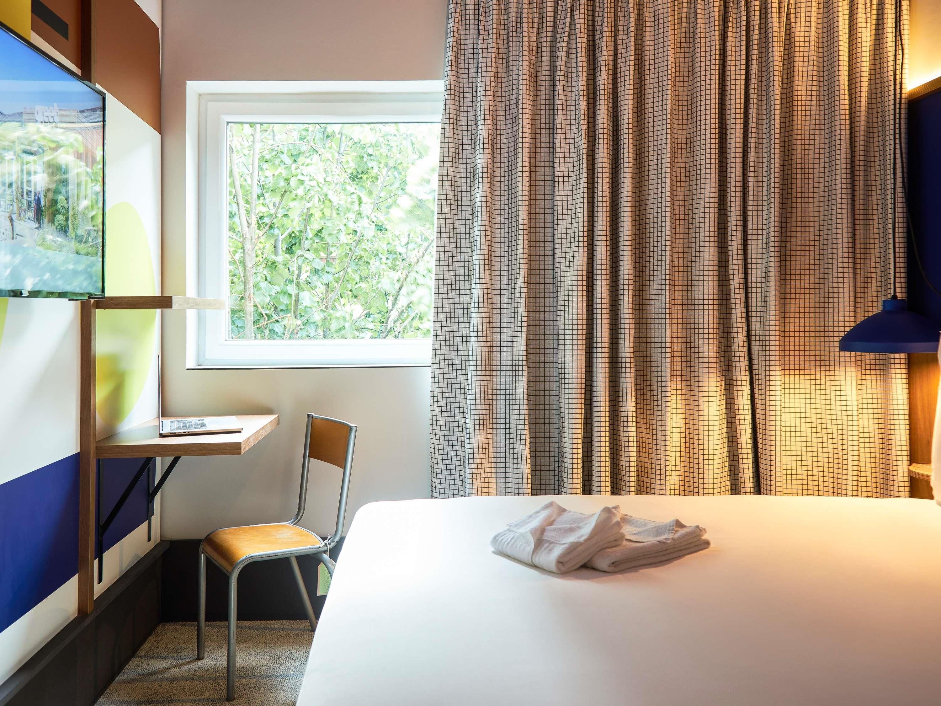 A hotel room with a bed, a desk, a chair, a window with a view of trees, and a curtain.