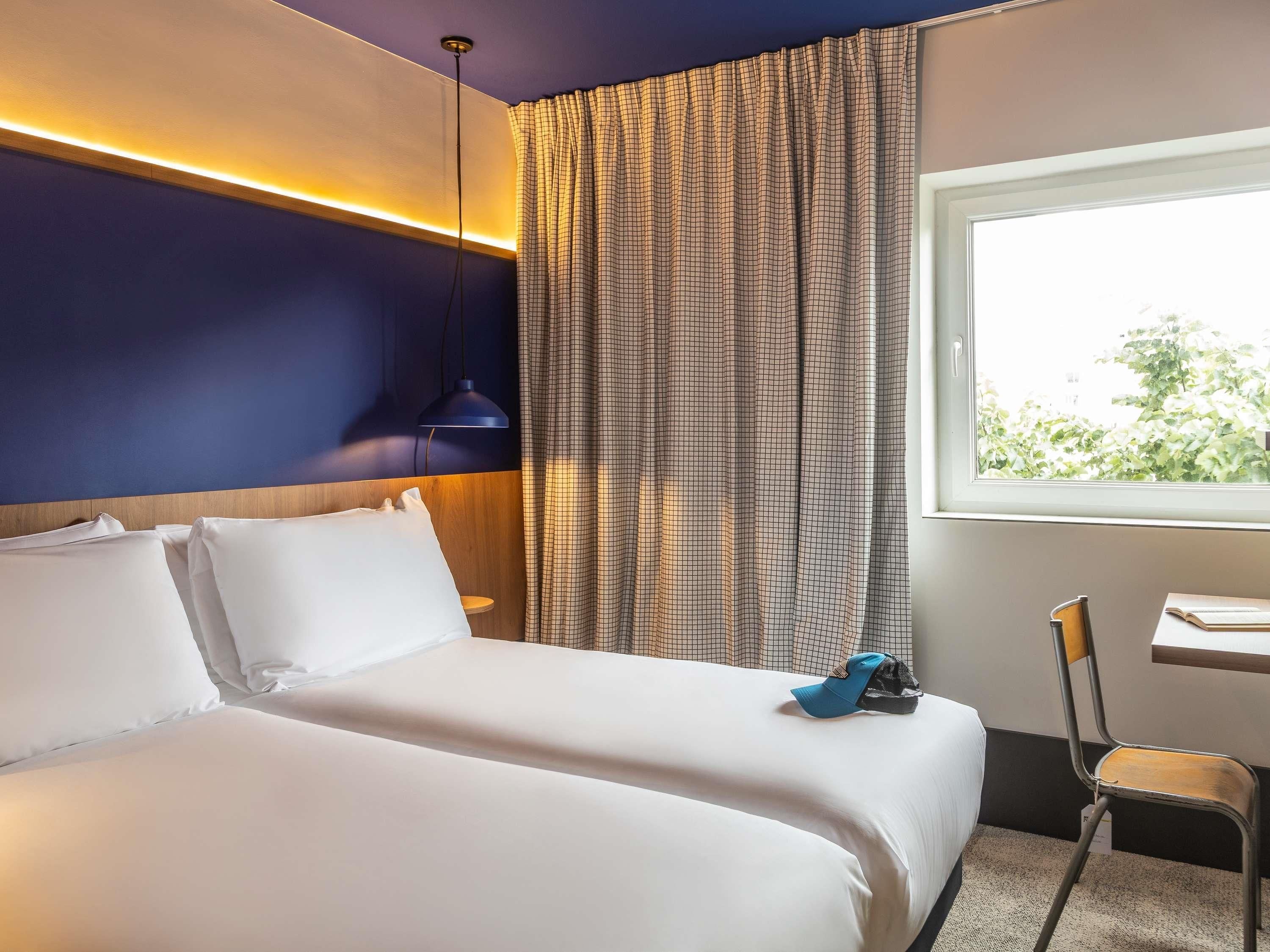 A hotel room with a bed, a desk, a chair, and a window with a view of greenery.