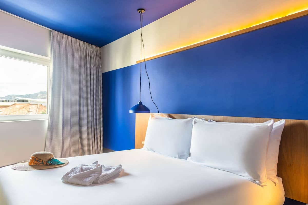 Une chambre d’hôtel avec un lit, une table de chevet, une lampe, une fenêtre avec des rideaux et une vue sur l’extérieur.