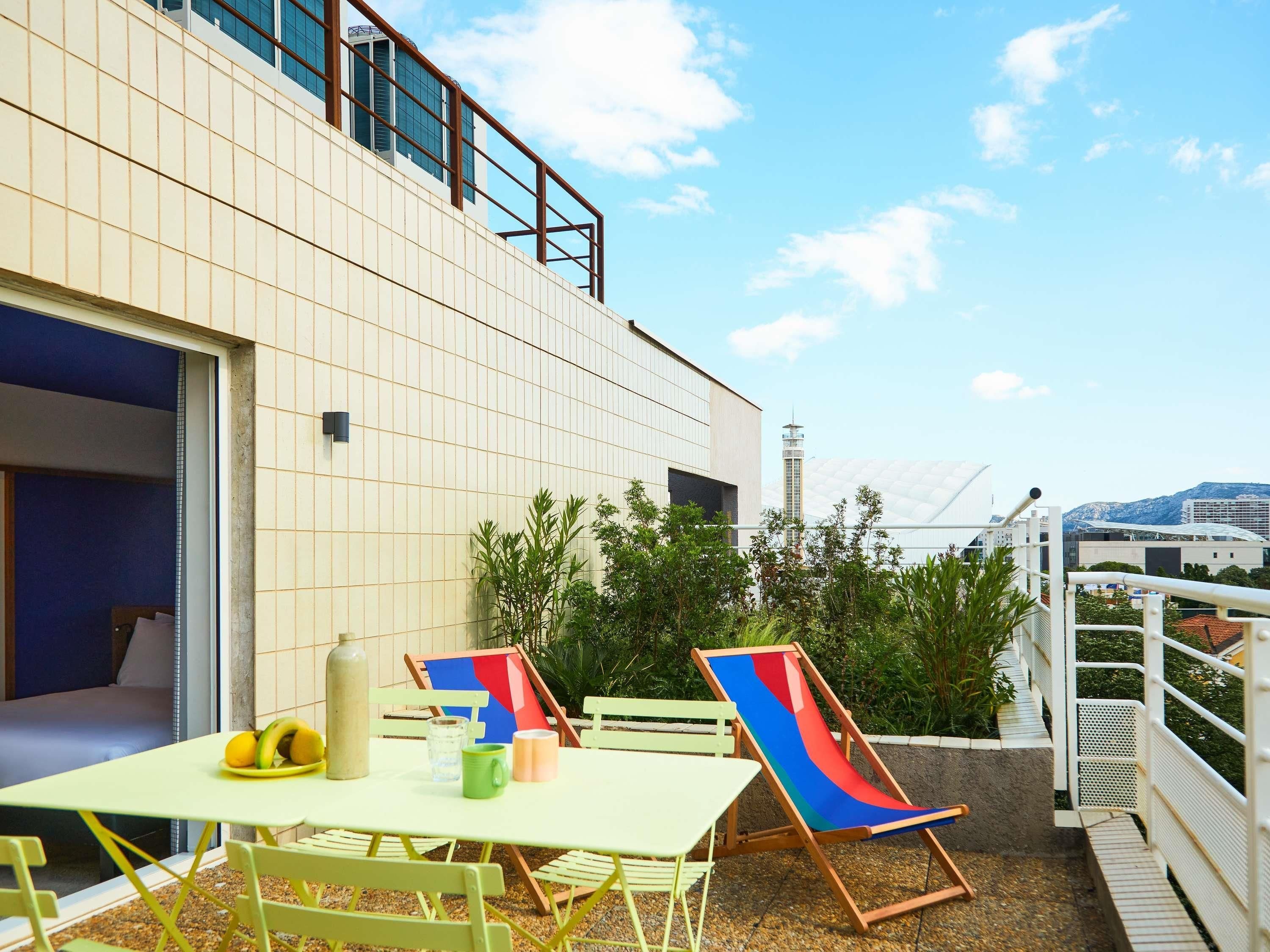 A modern building with a tiled facade, outdoor seating area with chairs and a table, and a view of a distant tower and mountains.