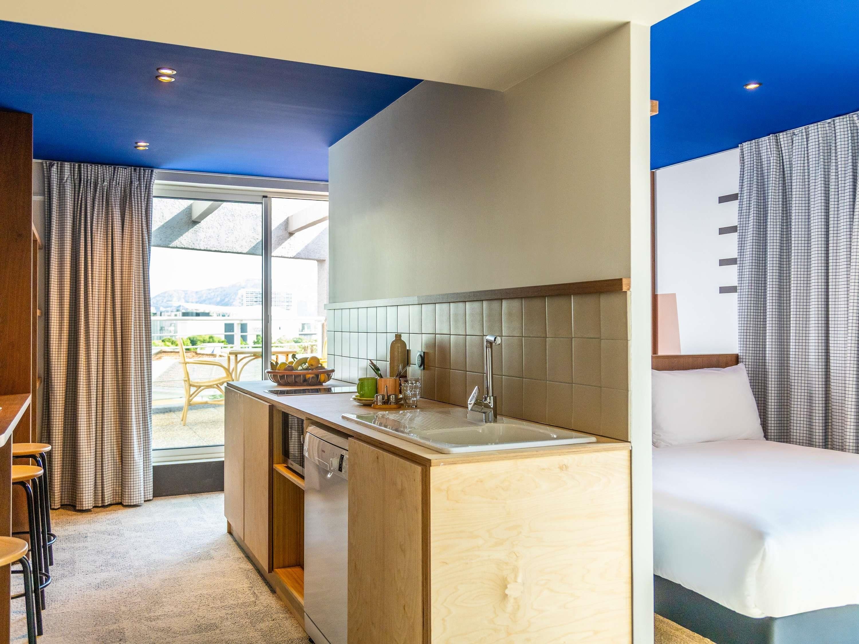 A modern kitchen with a dishwasher, a sink, and a countertop with various items. There is a dining area with chairs and a table, and a bed with a white mattress and pillows.