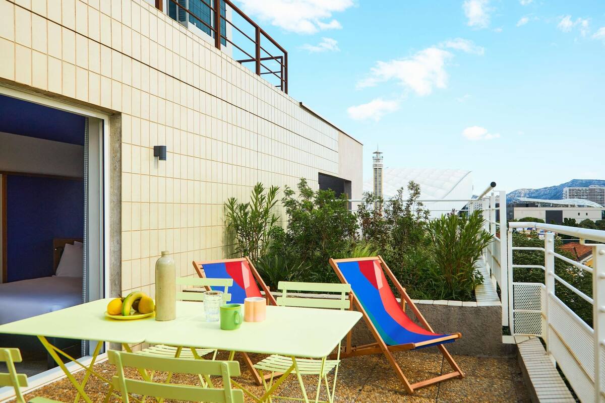 A modern building with a tiled facade, outdoor seating area with chairs and a table, and a view of a distant tower and mountains.