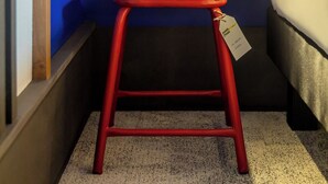Een klein houten krukje met een label, een bed met wit beddengoed en een wandlamp.