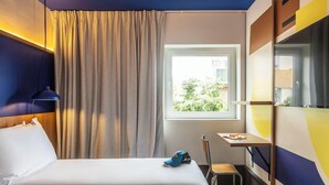 Una camera d'albergo con un letto, una scrivania, una sedia, una finestra con tende e vista su piante.
